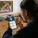 Fotorealistischer Schulterblick auf ein junges Mädchen, das am Schreibtisch ein Smartphone hält. Auf dem Display ist das Logo „Jugendschutz digital“ zu sehen, während an der weißen Wand im Hintergrund ein gerahmtes Familienfoto mit dem Titel „Profit auf Kosten der Kinder“ hängt.